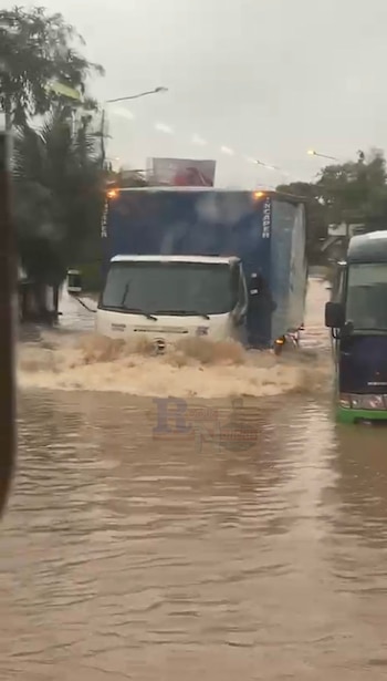 Un camión de caja blanco y azul oscuro atraviesa una calle severamente inundada con agua marrón turbia que salpica. Un autobús oscuro se ve a la derecha, también en el agua