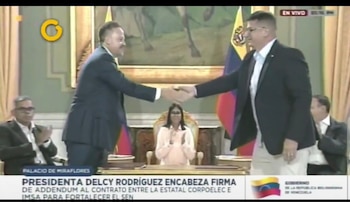 Dos hombres con traje se dan la mano frente a Delcy Rodríguez, quien permanece sentada entre banderas de Venezuela en una sala de reuniones