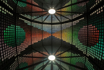 Vista interior de una cúpula abovedada con un patrón de rejilla y pequeños cuadrados de colores vibrantes, destacando dos luminarias circulares