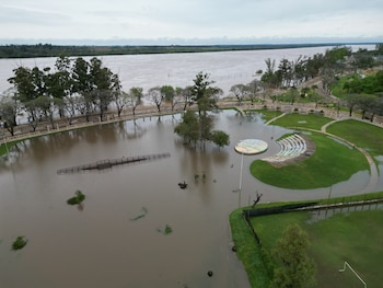 Fotografía aérea de un parque inundado por agua marrón junto a un río, con árboles, césped y un anfiteatro parcialmente sumergido