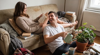 Hombre riega una planta con regadera verde junto a una mujer que lee un libro en un sofá, con libros y otras plantas en macetas en un hogar.