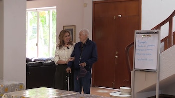Matilde Obregón y Mario Casillas, con bastón y gorra, de pie en un interior. Un pizarrón blanco con menú y una escalera de madera son visibles
