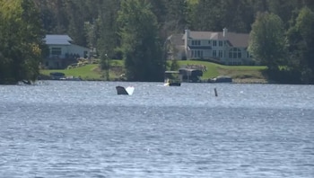 La cola de una avioneta sobresale de la superficie de un lago, con casas y árboles en la orilla de fondo