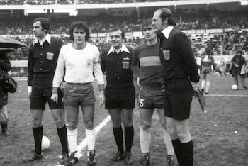 Cinco hombres parados en una cancha de fútbol: tres árbitros con uniforme oscuro y dos jugadores, uno con camiseta clara y otro con camiseta a rayas, junto a un balón