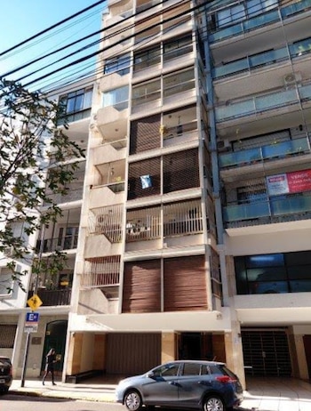 Edificio alto de apartamentos, balcones con persianas, ventanas, letrero rojo de "Vende", automóvil gris estacionado, persona en acera, señales de tránsito, cables