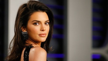 Primer plano de Kendall Jenner mirando por encima de su hombro derecho, luciendo cabello castaño oscuro, maquillaje natural, y un pendiente rectangular negro brillante