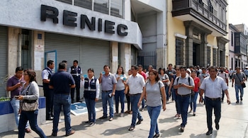 Personal de RENIEC con chalecos y documentos en mano en la entrada de un edificio, con personas caminando por la acera.