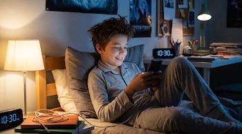 Un niño en pijama acostado en la cama usando un teléfono móvil. Se ven una lámpara de noche, un reloj digital, libros y un escritorio.