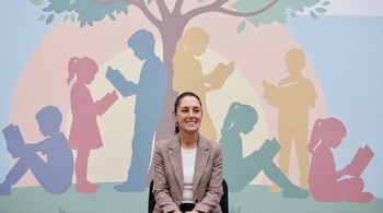 Claudia Sheinbaum, vestida con blazer a cuadros, sonríe sentada frente a un mural colorido con siluetas de niños leyendo bajo un árbol.
