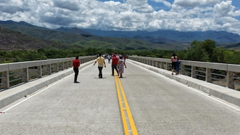 Vista frontal de un puente de concreto con varias personas caminando sobre él, rodeado de montañas verdes bajo un cielo nublado