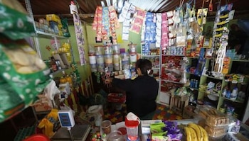 Interior de una bodega peruana con una mujer de espaldas atendiendo detrás de un mostrador, rodeada de estantes con productos y paquetes colgando del techo