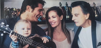 Dos hombres, una mujer y un bebé con una pequeña guitarra frente a un estante con libros y premios