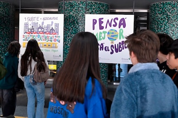 Varios jóvenes de espaldas miran dos carteles ilustrados sobre el uso del subte colocados frente a una columna de azulejos