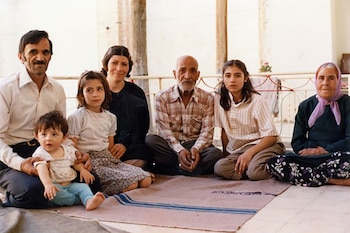 Siete personas, desde un bebé hasta ancianos, sentadas en el suelo sobre una manta beige con franjas azules, posan para un retrato familiar en un interior luminoso