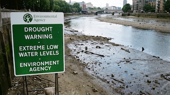 Cartel verde y blanco de la Agencia de Medio Ambiente que advierte sobre sequía y niveles de agua bajos, junto a la orilla expuesta de un río en una ciudad.