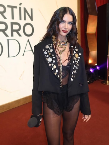 Zaira Nara de pie en una alfombra roja, luciendo un blazer negro corto decorado con medallones, lencería negra, pantimedias y una cadena dorada gruesa
