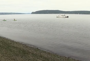 Vista aérea de un cuerpo de agua con un barco blanco, dos kayaks verdes con personas, una orilla rocosa y vegetación, y un horizonte boscoso bajo un cielo gris