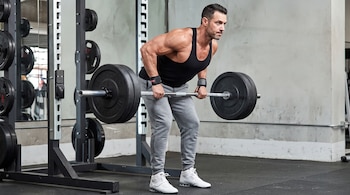 Hombre atlético realiza remo con barra cargada con discos en un gimnasio con rack de pesas y suelo de goma.