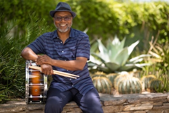 Hombre sentado con sombrero y gafas, sosteniendo baquetas y apoyado en un tambor. Fondo de vegetación con cactus y agave