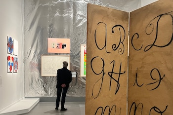 Un hombre de espaldas contempla cuadros colgados en una pared plateada, al lado de un biombo de madera con letras trazadas a mano