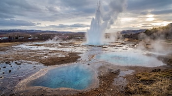Un géiser lanza un chorro de agua y vapor al aire cerca de pozas de agua azul en un valle rocoso bajo un cielo nublado.
