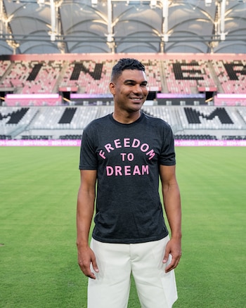 Un hombre de tez oscura sonríe en un campo de fútbol. Viste camiseta gris con texto rosado "FREEDOM TO DREAM" y shorts claros. Tribunas de estadio de fondo