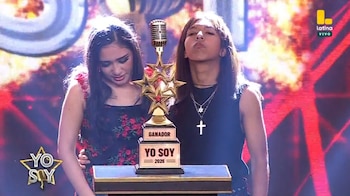 Dos personas permanecen de pie detrás de un trofeo con forma de micrófono y estrellas en un plató con pantallas iluminadas