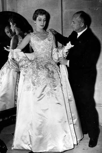 Una foto en blanco y negro muestra al diseñador Christian Dior ajustando el hombro del vestido de gala de su hermana Catherine, quien lleva un collar de perlas