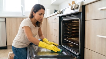 Joven mujer latina con guantes amarillos y pelo oscuro, limpiando el interior de un horno de cocina con una almohadilla verde en una cocina moderna.