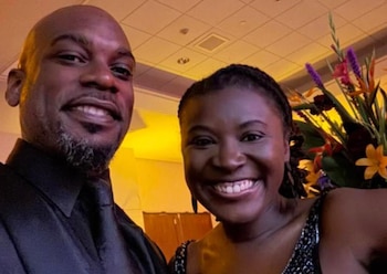 Andrew DeYoung y una mujer sonrientes posan para una foto en un interior, con iluminación cálida y un arreglo floral multicolor a la derecha