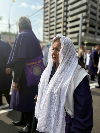 Una mujer con mantilla blanca y túnica morada camina apoyada en un bastón entre personas con hábitos morados en una calle urbana