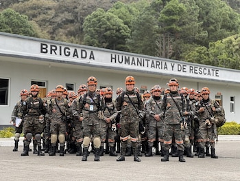 Grupo de personas con uniformes de camuflaje, cascos naranjas y equipo táctico. Edificio con letrero "BRIGADA HUMANITARIA Y DE RESCATE"