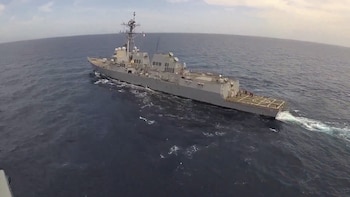 Vista aérea de un destructor de la Marina de EEUU navegando en el océano azul. El buque de guerra gris deja una estela blanca y tiene una cubierta de aterrizaje