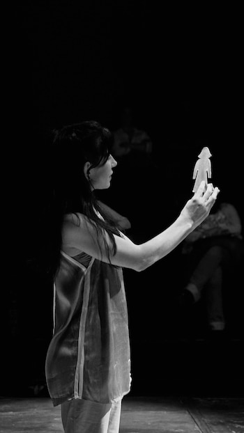 Imagen en blanco y negro de una mujer de perfil con cabello oscuro, sosteniendo en alto una figura de mujer recortada contra un fondo oscuro