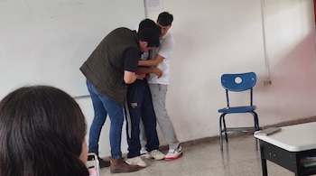 Tres jóvenes se abrazan junto a una pared blanca. Se ve una silla azul, un escritorio con un teléfono móvil y parte de una cabeza.