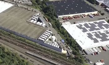 Vista aérea de varias naves industriales con paneles solares, camiones estacionados y vías de ferrocarril junto a áreas verdes
