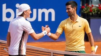 Dos tenistas, Ignacio Buse y Novak Djokovic, se estrechan las manos en una cancha de tierra batida. Buse lleva gorra blanca, Djokovic camiseta amarilla. Fondo azul.