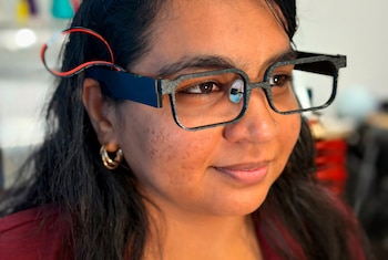 Una mujer de cabello oscuro lleva monturas de anteojos negros con cables rojos y negros conectados en la patilla derecha