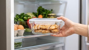 Una mano introduce un recipiente de vidrio con pollo cocido en rodajas en un refrigerador abierto que contiene huevos y verduras