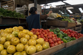 Maracuyás amarillos, tomates rojos y pimientos en cajas de madera ante una mujer que sostiene una balanza metálica.
