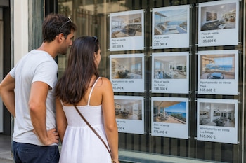 Un hombre y una mujer de espaldas observan varios carteles con fotos y precios en euros de pisos en venta pegados en un cristal.