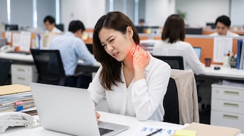 Mujer en oficina sentada frente a computadora portátil con expresión de dolor; una mano sujeta su cuello donde las cervicales están resaltadas en rojo.