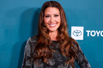 Primer plano de Shannon Elizabeth sonriendo, con cabello castaño ondulado suelto, maquillaje en los ojos y un vestido oscuro con detalles brillantes
