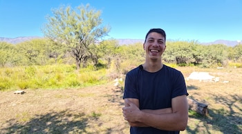 Un hombre con camiseta negra posa con los brazos cruzados en un terreno con vegetación, árboles y montañas al fondo.