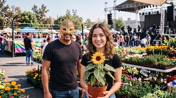 Una mujer sonriente sostiene un girasol en maceta junto a un hombre con máscara dorada en una feria al aire libre con puestos de flores.