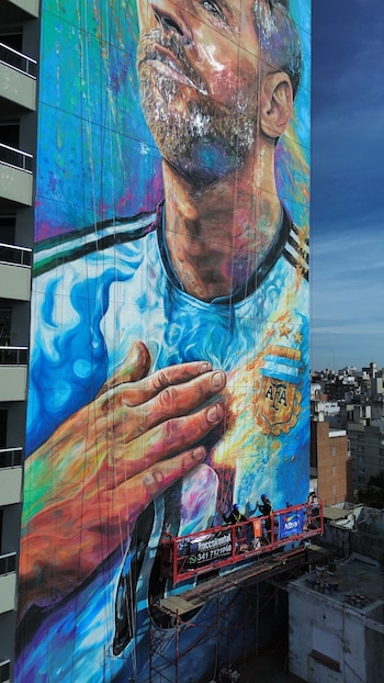 Mural gigante de Lionel Messi con camiseta de la selección argentina en la fachada de un edificio. Dos personas en plataforma elevadora trabajan frente a él