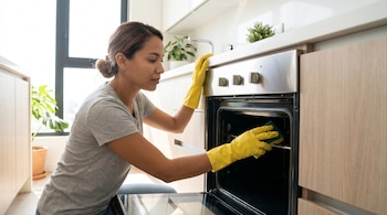 Una mujer latina joven, de piel clara y cabello oscuro, con guantes de goma amarillos, limpia el interior de un horno de cocina con un estropajo verde.