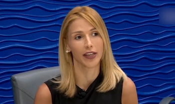 Una mujer rubia vestida de negro habla frente a una pared de color azul con relieve en forma de ondas