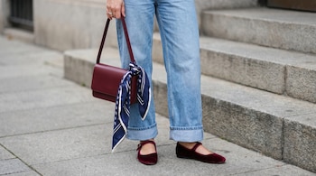 Primer plano de las piernas de una persona vistiendo jeans anchos, bailarinas de terciopelo burdeos y un bolso de hombro del mismo color con un pañuelo azul atado.