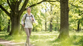 Una mujer de mediana edad con cabello corto y gris camina en un sendero de tierra en un parque, llevando una mochila y una botella de agua, rodeada de árboles y vegetación verde.